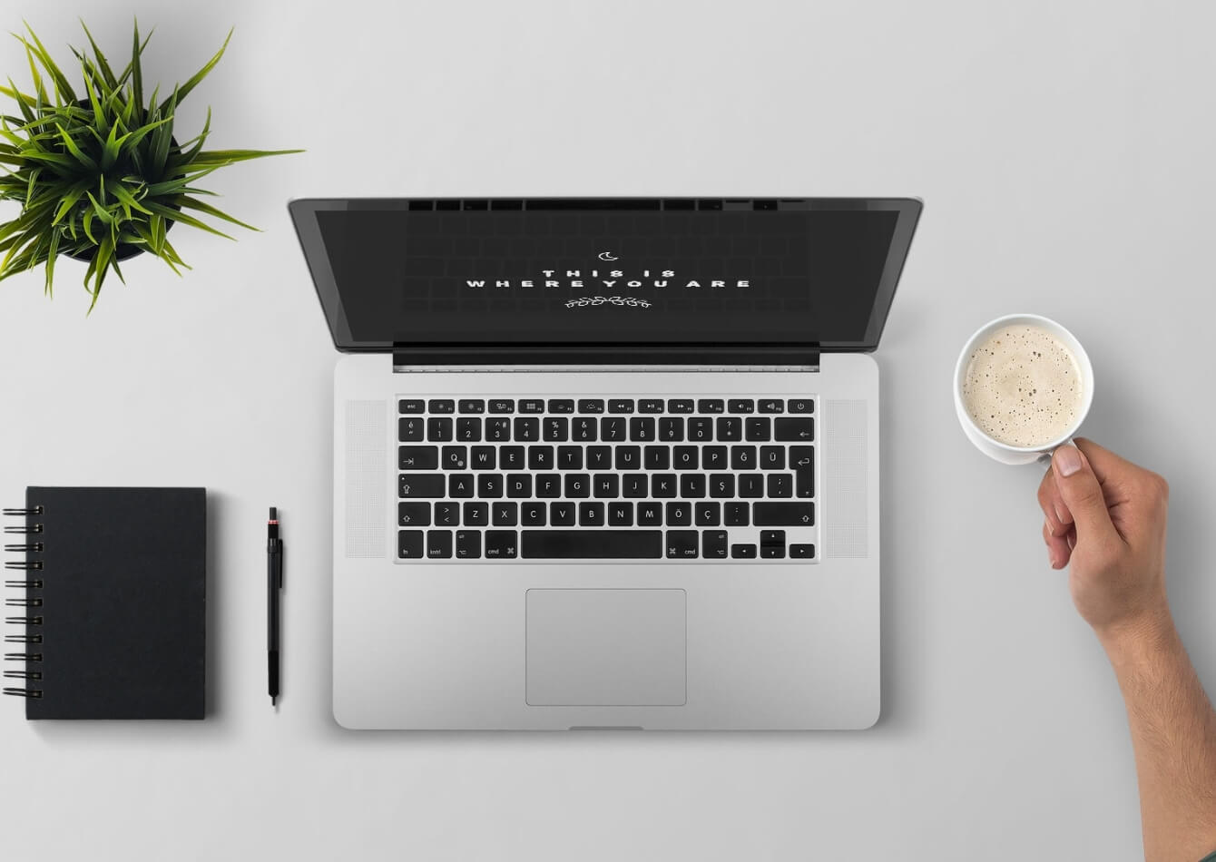 laptop, pen, notebook, plant on a table and a hand holding a coffee cup